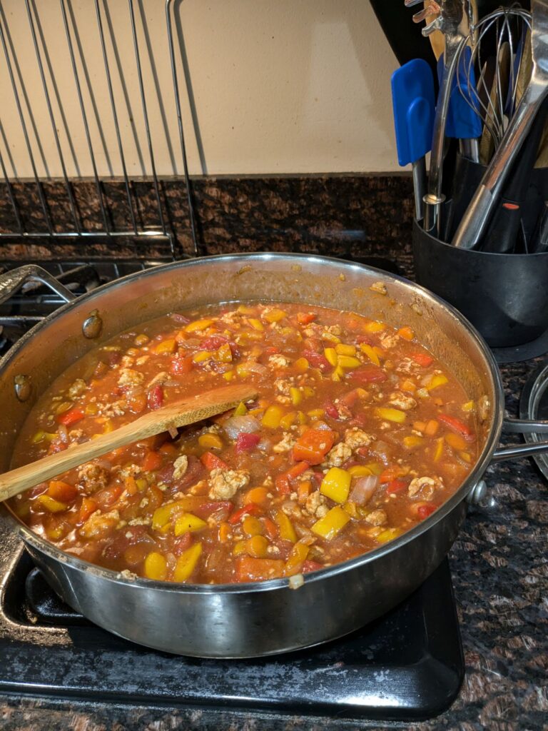 A pot of chili simmering in a metal pan on a stove. It is very red. There is a wooden spoon resting on the chili.